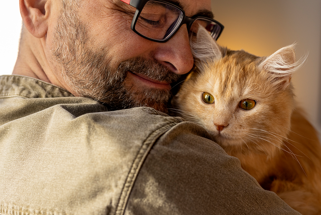 Homem sorridente segurando um gato laranja no colo, em um momento de carinho e conexão. O gato observa atentamente o ambiente ao redor.